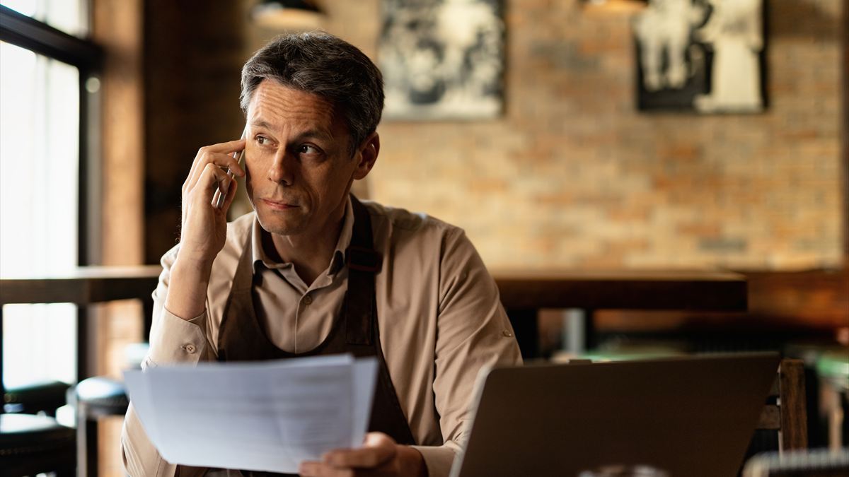 Dono de um pequeno restaurante ao telefone, com papéis na mão e o notebook aberto sobre a mesa
