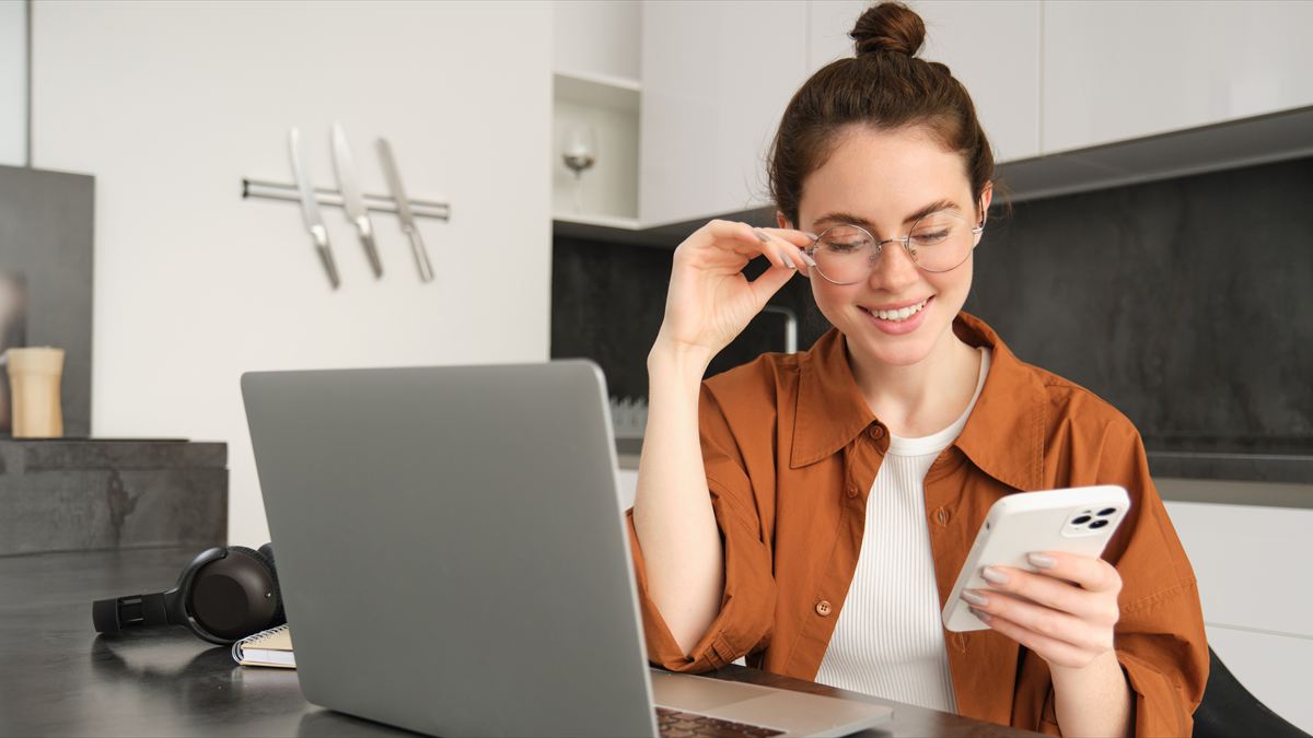Profissional sorrindo enquanto responde mensagens no celular, com o notebook aberto na bancada