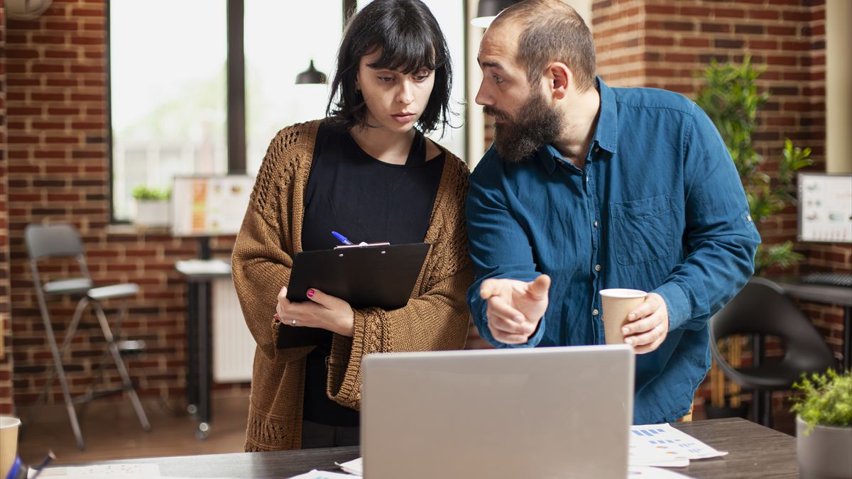 Duas pessoas conversando sobre um projeto em volta de um notebook, uma delas com uma prancheta
